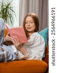 Teenager girl relaxing reading interesting book, turning pages smiling enjoying literature, taking a rest on comfortable couch. Happy child kid at home apartment living room lying on sofa. Vertical