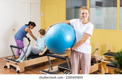 Teenager girl standing in gym with big blue fitness ball in hands and looking in camera. Pilates trainer helping youth boy to do exercises on reformer and use pilates ring in background.