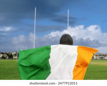 Teenager girl standing and holding National flag of Ireland. Selective focus. Tall goal posts and sport field for Irish sport in the background. Supporting hurling, camogie, rugby and Gaelic football - Powered by Shutterstock