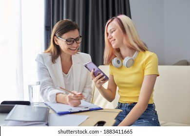 Teenager Girl Talking To School Psychologist, Smiling Student Sitting On The Couch In Office Of Social Worker. Professional Counselor Looks At Smartphone Screen With Student