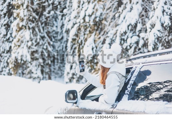 Teenager girl\
traveling looking out of car window and taking selfie on smartphone\
in winter snowy forest. Road trip and local travel concept. Happy\
child enjoying car\
ride.