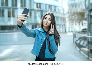Teenager girl in urban city take selfie picture by smartphone in public with fingers symbolize peace and mouth shaped as kissing duck face for followers at social media