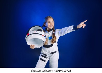 Teenager girl wearing astronaut costume and helmet standing sideways on dark blue background with copyspace. Cute girl in spacesuit pointing up at blank space for text.