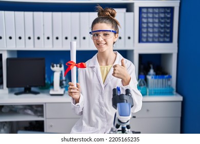 Teenager Girl Working At Scientist Laboratory Holding Degree Smiling Happy And Positive, Thumb Up Doing Excellent And Approval Sign 