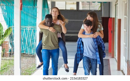 Teenager Group Of Multi Ethnic Friends At School Having Fun Piggybacking Outdoors In The Hallway.