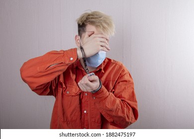 A Teenager In Handcuffs And A Medical Mask Holds His Head Against A Gray Background.Headache For Juvenile Delinquents In Quarantine, Criminal Liability. Members Of Youth Criminal Groups.