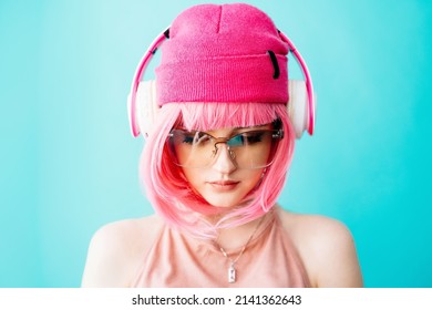 A Teenager In Headphones  And Glasseswith Pink Hair. Bright Extravagant Appearance. Modern Youth Culture. Portrait Of A Girl With A Short Haircut On A Blue Background. Mockup Friendly. 