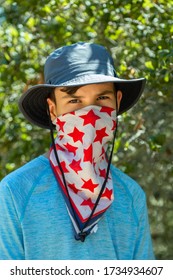 Teenager Hikes In The Park And Covers His Face With A Bandana Mask With Red Stars. Summer, Green Trees Background. The Boy Is Wear A Cloth Face Covering To Help Prevent The Spread Of COVID-19
