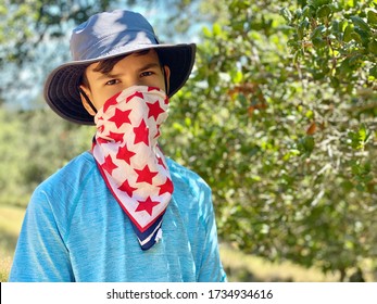 Teenager Hikes In The Park And Covers His Face With A Bandana Mask With Red Stars. Summer, Green Trees Background. The Boy Is Wear A Cloth Face Covering To Help Prevent The Spread Of COVID-19
