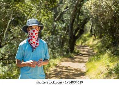 Teenager Hikes In The Park And Covers His Face With A Bandana Mask With Red Stars. Summer, Green Trees Background. The Boy Is Wear A Cloth Face Covering To Help Prevent The Spread Of COVID-19