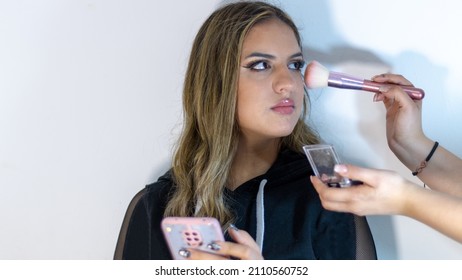 Teenager holding her phone, while  her friend is doing makeup to her