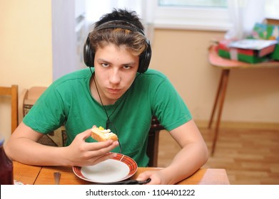 The Teenager Listens To Music In The Headphones, Sits In The Kitchen And Eats A Sandwich. He Has Angry Face Because He Is Prevented From Eating And Calls Him To Clean His Room