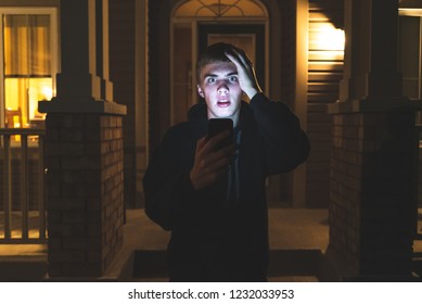 Teenager Looking At Something Shocking On His Mobile Phone. He Is Standing In Front Of His House At Night.