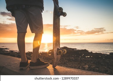 Teenager Looking At Sunset On The Horizon Holding A Skateboard In Hand. Teen Wearing Plaid Shirt, Shorts And Black Sneakers The Sun Sets, Golden Reflections On The Shore Of The Ocean. Back Close Up