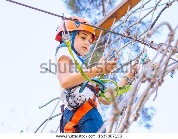 Teenager in orange helmet climbing in trees on
forest adventure park. Girl walk on rope cables and high suspension
bridge in adventure summer city park. Extreme sport equipment
helmet and carabiner.