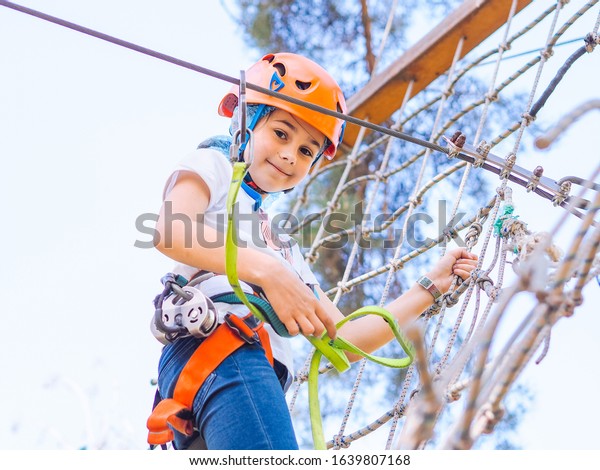 Teenager in orange helmet climbing in trees on\
forest adventure park. Girl walk on rope cables and high suspension\
bridge in adventure summer city park. Extreme sport equipment\
helmet and carabiner.
