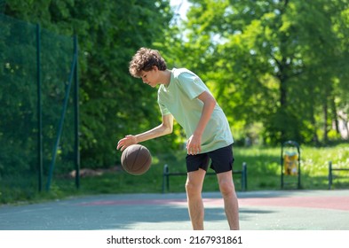 Teenager Playing Basketball Smiling, Cheerful. Fit Boy With Basketball Ball Outdoors. Kid Dribbling The Ball At The Court