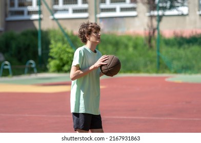 Teenager Playing Basketball Smiling, Cheerful. Fit Boy With Basketball Ball Outdoors. Kid Dribbling The Ball At The Court