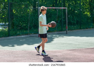 Teenager Playing Basketball Smiling, Cheerful. Fit Boy With Basketball Ball Outdoors. Kid Dribbling The Ball At The Court