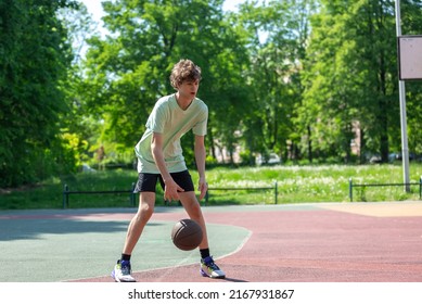 Teenager Playing Basketball Smiling, Cheerful. Fit Boy With Basketball Ball Outdoors. Kid Dribbling The Ball At The Court