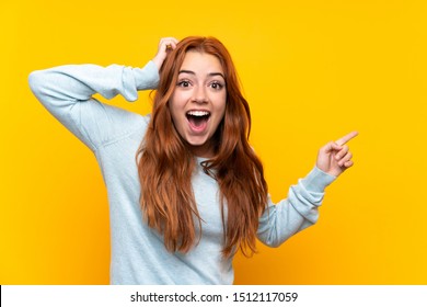 Teenager redhead girl over isolated yellow background surprised and pointing finger to the side