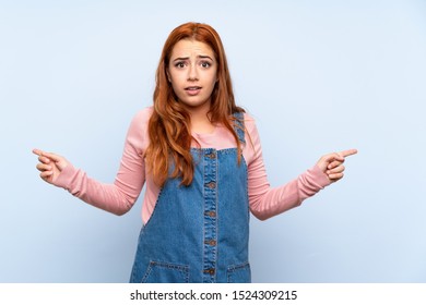 Teenager redhead girl with overalls over isolated blue background pointing to the laterals having doubts