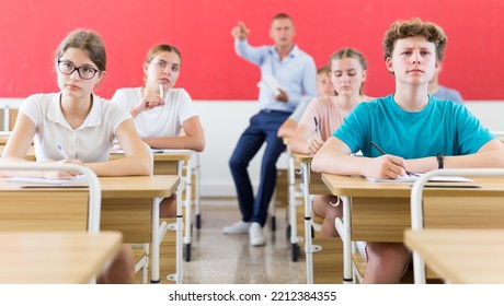 Teenager Sitting In Class Room. Male Teacher Explaining Something To Them.