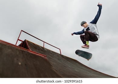 Teenager skateboarder in a cap doing a trick jump on a half pipe on a cloudy sky background