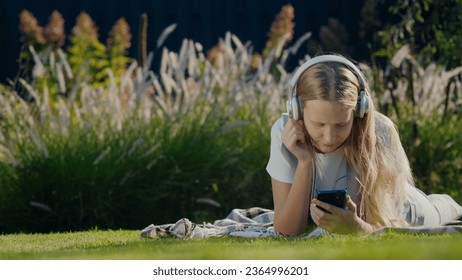 A teenager with a smartphone and headphones lies on the grass in the park. - Shutterstock ID 2364996201