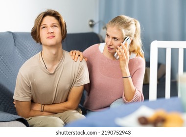 Teenager With Stomach Ache Sitting On Couch At Home And Holding His Stomach With Hands While Worried Mom Calling Doctor On Phone