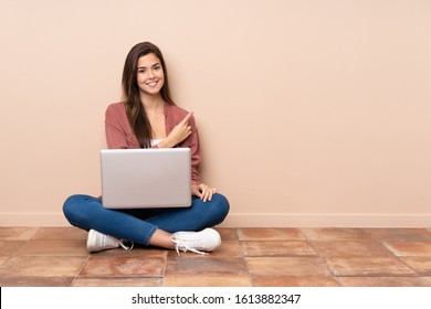 Teenager Student Girl Sitting On The Floor With A Laptop Pointing To The Side To Present A Product