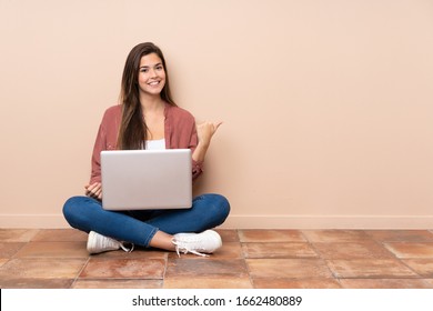 Teenager Student Girl Sitting On The Floor With A Laptop Pointing To The Side To Present A Product