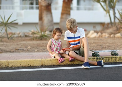 Teenager In T Shirt And Little Girl Looking On Mobile Phone At Urban Street. Siblings Spends Free Time In The City Park. Children Enjoys Sunny Day Outdoors With Skateboard. Hobby, Sport For Kids.