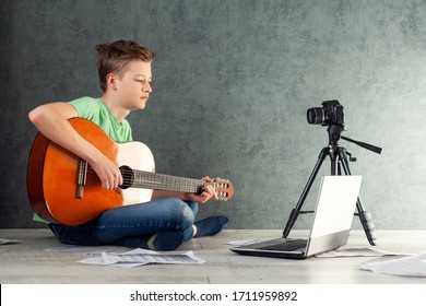 Teenager Takes Off The Vlog Of A Game On A Guitarat Home On The Camera. Young Videobloger Teen Boy Is Learn To Play The Guitar Online In Living Room, Resting At Home.