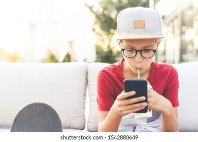 Teenager Watching Video On Smartphone Drinking A Cold Beverage. Young Boy Drinks Smoothie Sitting Outside At The Bar Using Cell Phone. Teen Using Mobile Phone Reading Friends Chat. New Tech Concept