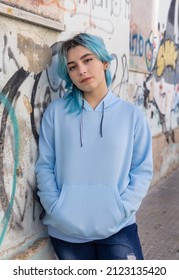 Teenager Wearing Light Blue Oversize Hoodie And Jeans Looking Into The Camera. Blue Haired Teen Girl Stays Outdoor Against Graffiti Wall. Clothing Mockup