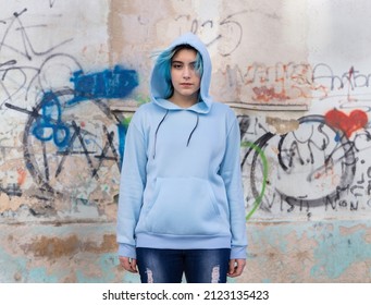 Teenager Wearing Light Blue Oversize Hoodie And Jeans Looking Into The Camera. Blue Haired Teen Girl Stays Outdoor Against Graffiti Wall. Clothing Mockup