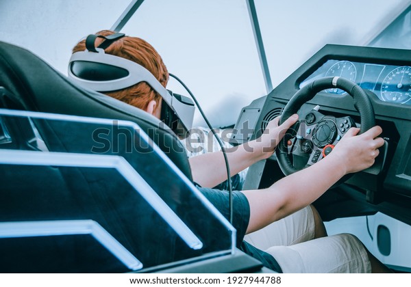 A teenager\
wearing virtual reality glasses sits in a moving interactive chair\
and holds the steering wheel. Virtual games. Virtual car driving.\
Artificial reality.