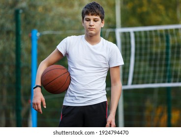 Teenager in a white t-shirt with orange basketball ball outside. Cute young boy plays basketball on the street playground in summer. Hobby, active lifestyle, sports activity for kids.