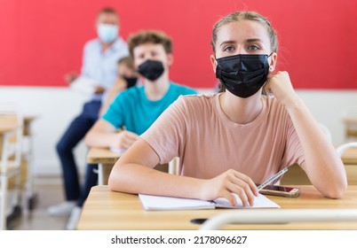 Teenagers In Face Masks Sitting In Class Room. Male Teacher Explaining Something To Them.