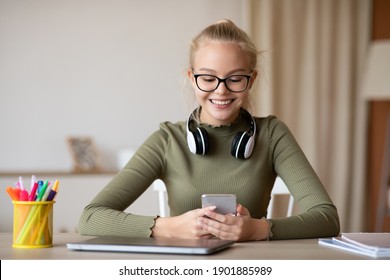 Teenagers Gadget Addiction Concept. Cheerful Teen Girl Using Mobile Phone And Smiling, Sitting At Desk, Relaxing After Doing Homework, Chatting With Friends, Home Interior, Copy Space