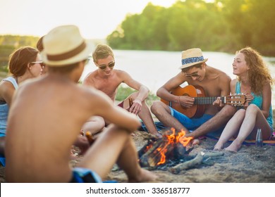 Teenagers Having Fun At The Beach. They Are Sitting In The Sand In Circle Around A Camp Fire, Wearing Short Pants, Sunglasses And Straw Hats. They Are Singing And A Boy Is Playing Guitar