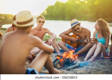 Teenagers Having Fun At The Beach. They Are Sitting In The Sand In Circle Around A Camp Fire, Wearing Short Pants, Sunglasses And Straw Hats. They Are Singing And A Boy Is Playing Guitar