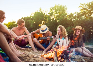 Teenagers having fun at the beach. They are sitting in the sand in circle around a camp fire, wearing short pants, sunglasses and straw hats. They are singing and a boy is playing guitar - Powered by Shutterstock