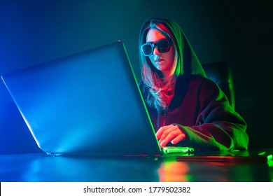 Teenagers Hobbies. A Teenager Watches A Video On The Internet. A Girl With Dark Glasses Is Sitting At A Laptop. The Girl At The Computer Is Illuminated By Colored Neon Lights.