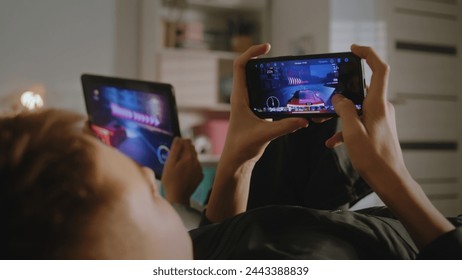 Teenagers lie on bed in their room and play video games on digital devices. Girl uses tablet, boy uses phone to play together. Sister spending leisure time with her brother. Home with cozy interior. - Powered by Shutterstock