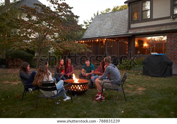 Teenagers sit talking around a fire pit in a garden\
at dusk