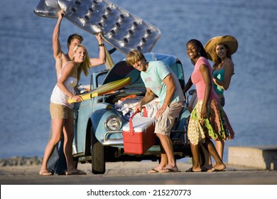 Teenagers Standing Beside Car Near Water, Carrying Cool Box And Inflatable, Smiling