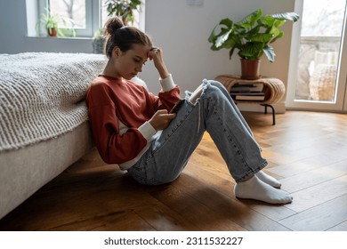 Teens and cyberbullying. Upset teen girl sitting on floor near bed using smartphone at home, scrolling social media. Child spending too much time on phone. Teenagers and gadget addiction - Powered by Shutterstock