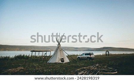 A teepee stands on the lakeshore next to a car, surrounded by windblown grass and the tranquil waters of the natural landscape